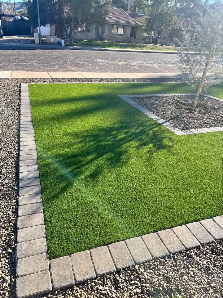A neatly manicured, vibrant green lawn with a smooth, curving edge meets a light gray concrete sidewalk and a row of red bricks. The background features shrubbery and a brick wall of an Arizona house, showcasing the artistry of artificial grass installations in Deer Valley.