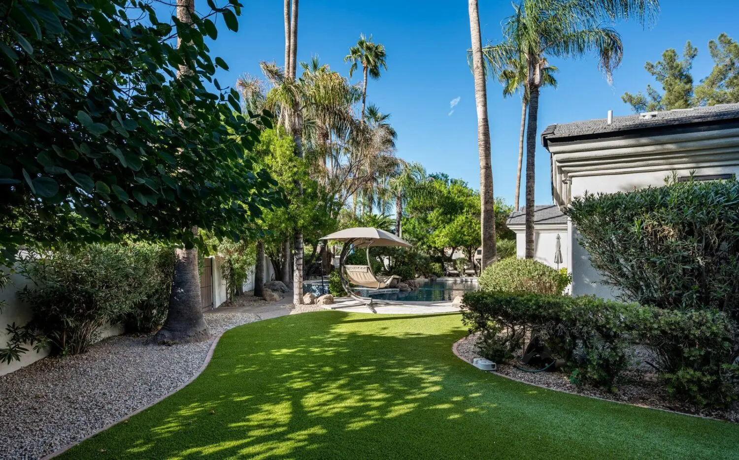 A neatly manicured, lush green lawn, thanks to an artificial grass installation, is intersected by a clean, curved brick border meeting a smooth concrete sidewalk. A house and plants enhance the neat and organized exterior setting, perfect for those seeking low maintenance lawns in Glendale AZ.