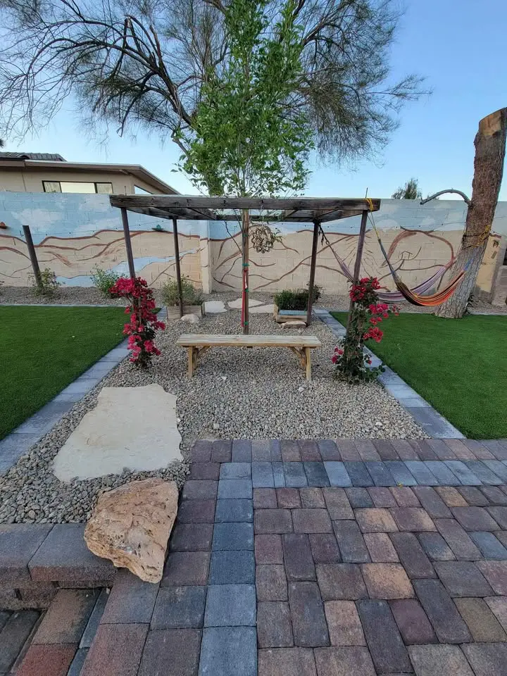 A small house with beige walls and brown trim sits in Deer Valley, surrounded by neatly trimmed bushes and a tree. The Arizona yard features landscaped rocks and artificial grass beneath the clear blue sky. A sidewalk borders the garden area.
