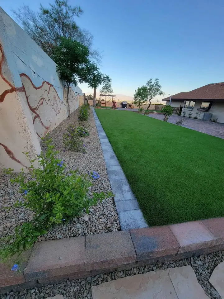 A small house with beige walls and brown trim sits in Deer Valley, surrounded by neatly trimmed bushes and a tree. The Arizona yard features landscaped rocks and artificial grass beneath the clear blue sky. A sidewalk borders the garden area.